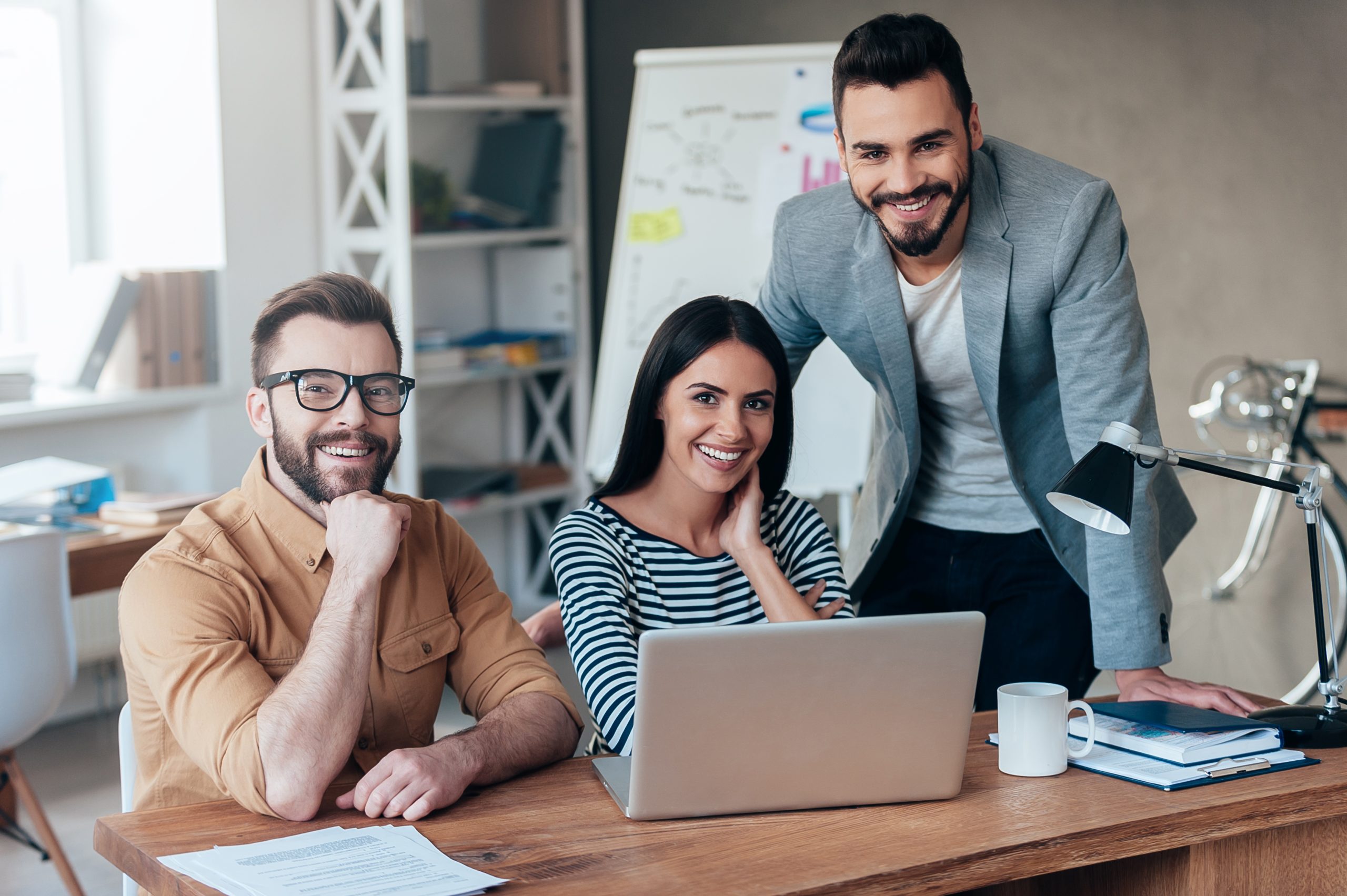 Successful team. Three confident business people in smart casual wear looking at camera and smiling while sitting at the desk in office