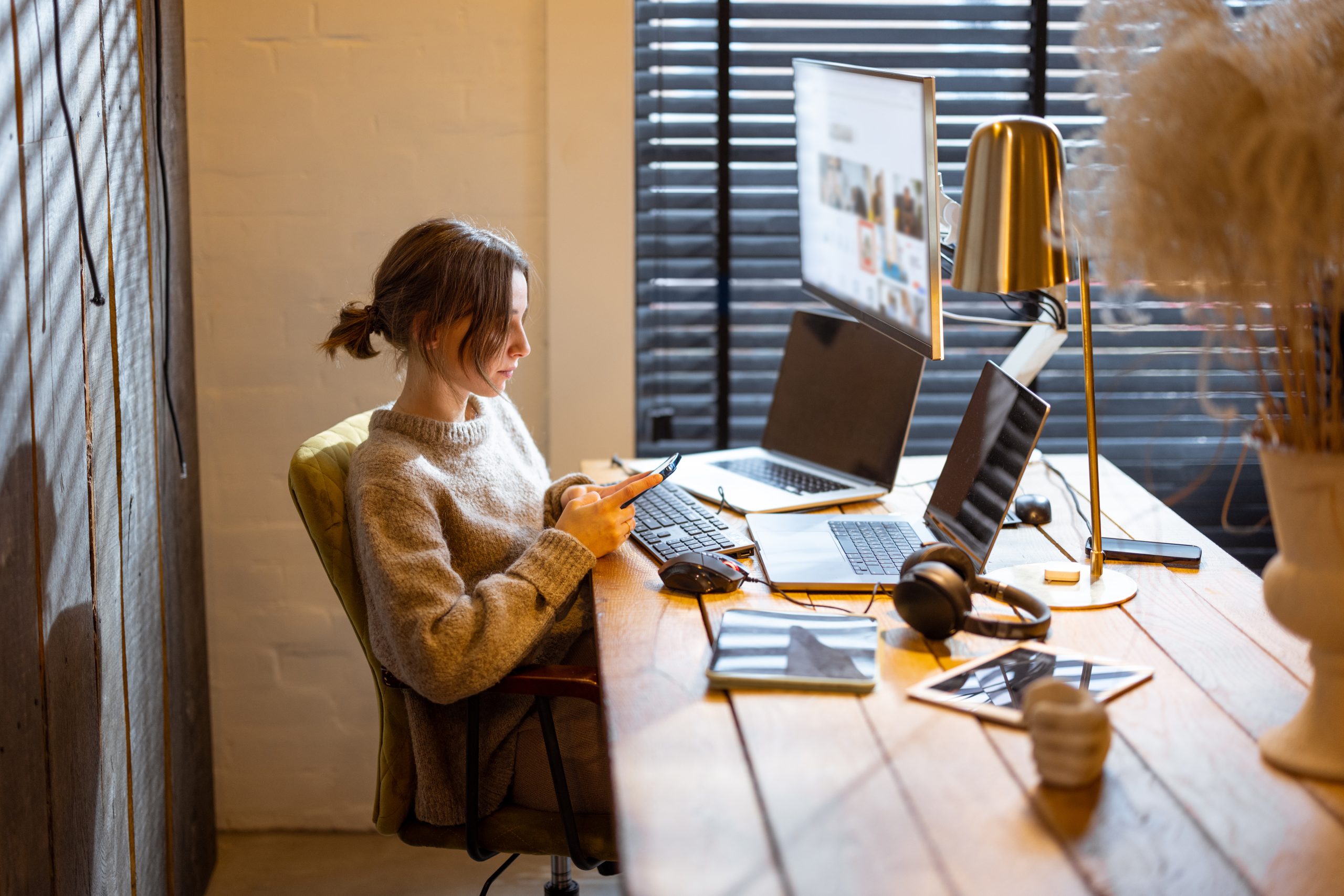 Young woman using phone while sitting at workplace with laptop and desktop computers at cozy home office interior. Concept of freelance and remote work at home. Light from the blinds on the window